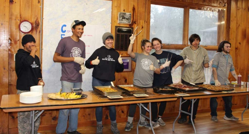 A group of people stand behind a long table lined with food, appearing to be preparing to serve during the family seminar of an outward bound intercept course.